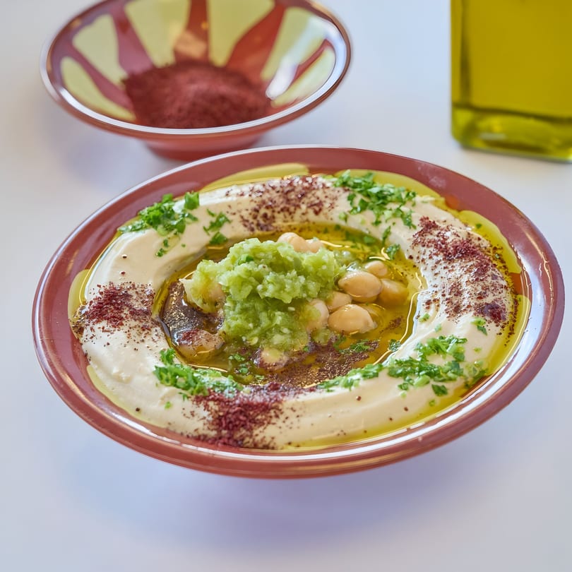 Creamy hummus topped with green relish, chickpeas, olive oil, sumac, and parsley, with a spice bowl.